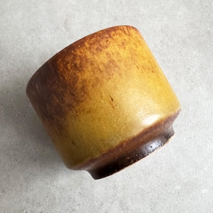 Könnte beinhalten: Eine handgefertigte Keramiktasse mit einer einzigartigen, erdigen Ästhetik. Die Tasse hat einen Farbverlauf, der von einem tiefbraunen Boden zu einem warmen gelbbraunen Rand übergeht. Das Design ist einfach und funktional, für Getränke.