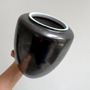 May include: A dark gray ceramic vase with a rounded body and a slightly flared rim. The rim is accented with a light blue band. The vase is held by a person's hand.