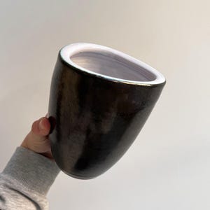 Può includere: Una tazza in ceramica scura e lucida con una forma asimmetrica unica. L'interno è bianco e il bordo è accentuato da una sottile fascia dorata. La tazza è tenuta su uno sfondo semplice.
