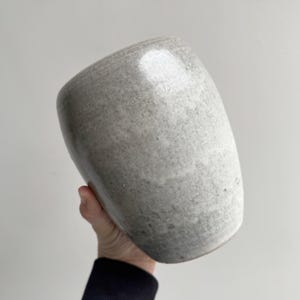Peut inclure: Un vase en céramique gris moucheté de forme arrondie et asymétrique. Le vase est tenu dans la main d'une personne, mettant en valeur sa surface lisse et texturée. La couleur neutre et la forme unique suggèrent un design moderne et minimaliste.
