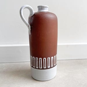 May include: A tall, brown ceramic vase with a white handle and a white base. The vase has a cylindrical shape with a decorative white rectangular pattern around the base. The handle is curved and attached to the top of the vase.