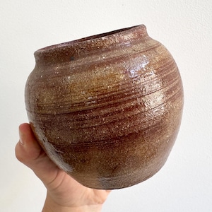May include: A small, round, brown ceramic pot with a glossy, textured finish. The vessel features horizontal ridges along its body, a wide opening at the top, and a mottled surface with variations in color ranging from dark brown to lighter tan tones.