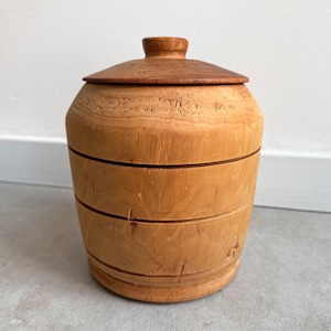 Puede incluir: Un tarro de madera marrón claro con tapa. El tarro tiene forma redondeada con bandas horizontales y un pequeño pomo en la tapa. La tapa es de un tono marrón más oscuro que el cuerpo del tarro. El tarro probablemente se usa para almacenamiento.