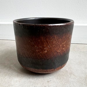 Könnte beinhalten: Ein brauner und schwarzer Keramiktopf mit strukturierter Oberfläche. Der Topf hat eine breite, runde Öffnung und einen leicht ausgestellten Rand.