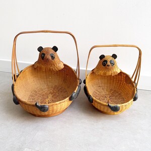 May include: Two round woven baskets with arched handles, featuring a panda bear head and black painted paws attached to the exterior. The baskets are made of light brown natural fibres with dark accents on the ears, eyes, and feet of the panda.
