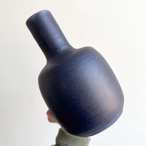 May include: A dark blue ceramic vase with a rounded body and a slender neck. The vase has a matte finish and is held against a neutral background. The design is simple and modern, suitable for home decor.