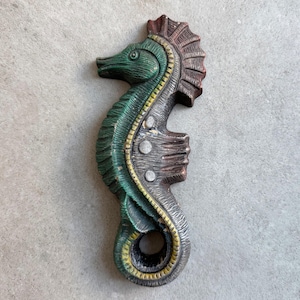May include: A carved seahorse sculpture with a detailed design. The seahorse is painted in shades of green, brown, and gray, with yellow accents. The sculpture has a textured surface and is likely made of wood or a similar material.