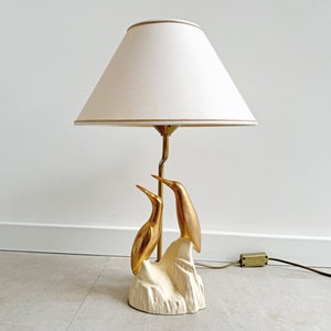 May include: A table lamp with a white lampshade and a gold bird sculpture base. The birds are perched on a white base.