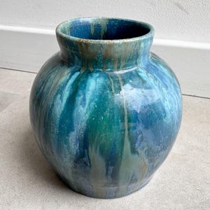Op de afbeelding: Een ronde keramische vaas met een brede opening en een korte hals. Het oppervlak heeft een glanzend glazuur met gemengde tinten diepblauw, turkoois en groenblauw, gekenmerkt door verticale druippatronen die een gelaagd effect creëren.