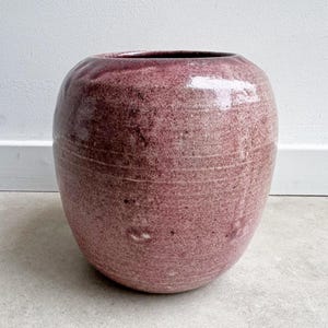 May include: A large, round, ceramic vase in a mottled, dusty rose color. The vase has a wide body and a slightly narrower opening. Horizontal lines encircle the body of the vase. The interior is a darker shade of burgundy.