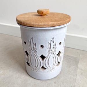 Op de afbeelding: Een witte keramische uienhouder met een houten deksel. De container heeft uitgesneden uienontwerpen en kleine ventilatiegaten. Het deksel heeft een kleine houten knop. Een keukenopbergartikel.