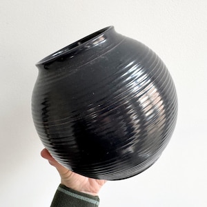 Pode incluir: Um vaso esférico preto de cerâmica com uma superfície texturizada e canelada. O vaso tem uma abertura larga e um pescoço ligeiramente curvo. A cor escura e a textura única conferem-lhe uma estética moderna e minimalista. Este vaso decorativo é adequado para exibir flores ou como peça de arte.