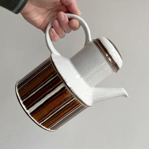 Peut inclure: Théière en céramique blanche et marron. Le corps présente des rayures verticales marron, le dessus et la poignée sont blancs. La théière est tenue en biais. Mesures en centimètres.