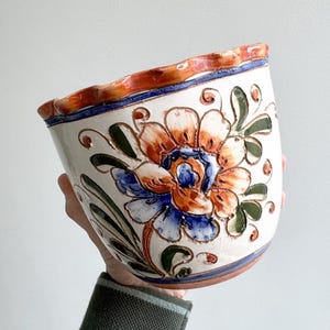 Op de afbeelding: Een handbeschilderde keramische bloempot met een bloemenontwerp. De bloempot heeft een grote oranje en blauwe bloem met groene bladeren op een witte achtergrond. De rand heeft een golvende vorm met oranje en blauwe accenten.
