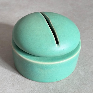 Könnte beinhalten: Eine tealfarbene Keramikdose mit Deckel und einem Schlitz oben. Die Dose ist rund und hat eine glatte, matte Oberfläche.