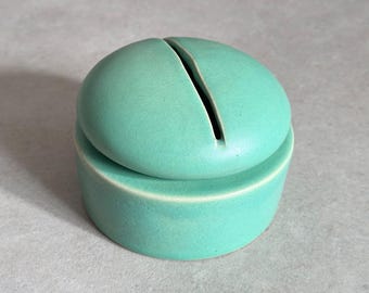 May include: A teal ceramic lidded jar with a slit in the top. The jar is round and has a smooth, matte finish.