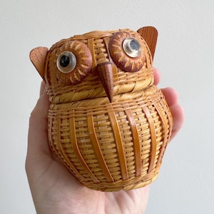 May include: A woven wicker owl-shaped container with a lid. The owl has large, round eyes with clear centers, a wooden beak, and small wooden ear-like accents. The container is held in a hand against a plain background.