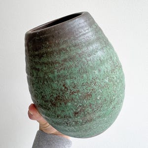Può includere: Un vaso in ceramica arrotondato con una superficie ruvida caratterizzata da uno smalto maculato verde e marrone scuro. Il contenitore presenta un'apertura ampia e leggermente inclinata sulla parte superiore e una base affusolata.
