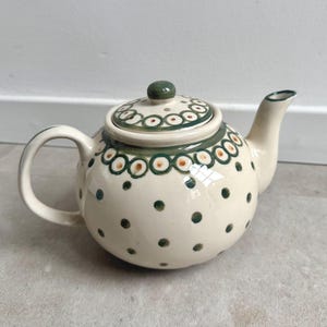 Op de afbeelding: Crème keramische theepot met een afgeronde body, gebogen handvat en tuit. Versierd met groene en oranje cirkelvormige patronen rond de rand en het deksel, en groene stippen. Het deksel heeft een groene knop. Ongeveer 20 cm lang.