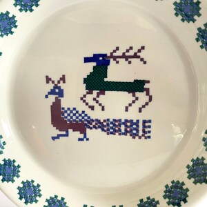 Pu&ograve; includere: Un piatto in ceramica bianca con un bordo blu e verde. Al centro del piatto, un design stilizzato di pavone e cervo in tonalit&agrave; di blu, verde e viola.