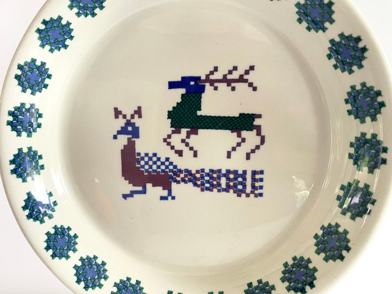 Pu&ograve; includere: Un piatto in ceramica bianca con un bordo blu e verde. Al centro del piatto, un design stilizzato di pavone e cervo in tonalit&agrave; di blu, verde e viola.