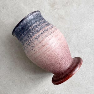May include: A handcrafted ceramic vase with a unique color gradient. The top rim is dark blue, transitioning to pink and then a deep red base. The vase has a textured surface and a flared base.