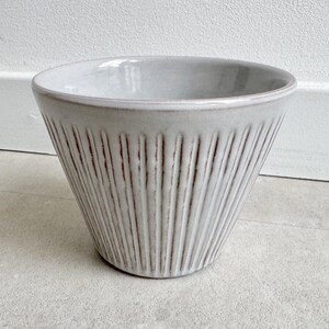 May include: A light grey ceramic bowl with a flared shape. The bowl has vertical, textured lines and a slightly darker rim. The interior is smooth and the bowl sits on a light grey surface.