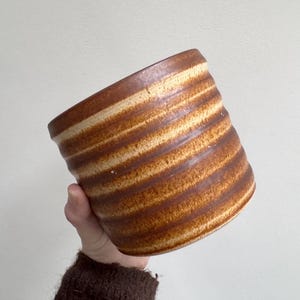 May include: A handmade ceramic pot with a cylindrical shape. The pot features horizontal stripes in shades of brown and beige. The pot is held by a person.