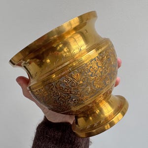 Peut inclure: Un bol en laiton doré avec un motif floral orné gravé au milieu. Le bol a un bord large et évasé et une base en piédestal. Le bol est tenu dans la main d'une personne.