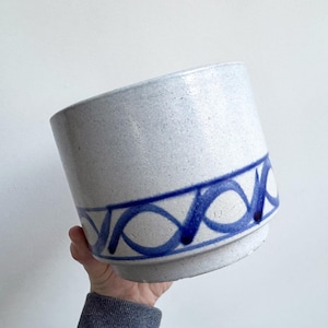 Può includere: Un vaso di ceramica blu chiaro maculato con un motivo a onde blu intorno alla parte superiore. Il vaso cilindrico ha una base leggermente svasata ed è tenuto da una persona.