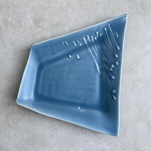 Puede incluir: Un plato de cerámica trapezoidal con un esmalte azul polvoriento y liso. La superficie presenta un patrón decorativo de líneas blancas finas en relieve y pequeñas gotas blancas redondeadas a lo largo del borde superior derecho.