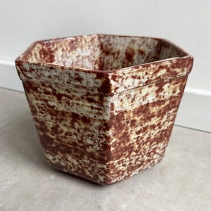 May include: A square ceramic plant pot with a textured surface. The pot has a brown and cream speckled pattern. The plant pot is empty and ready for planting. The pot is a decorative item for the home.