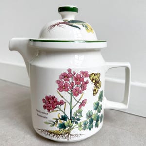 Könnte beinhalten: Eine weiße Keramik-Teekanne mit Deckel, verziert mit einer botanischen Illustration von rosa Cardamine pratensis Blüten, grünen Blättern und einem gelben Schmetterling. Die Teekanne hat einen dunkelgrünen Rand am Deckel und einen eckigen Henkel.