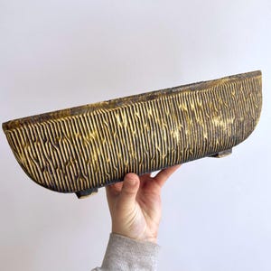Pode incluir: Um vaso de cerâmica alongado em forma de barco com um exterior texturizado de nervuras verticais. A superfície apresenta uma base cinzento-carvão com um acabamento dourado metálico desgastado nos relevos, apoiado sobre pequenos pés.
