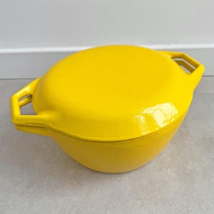 Könnte beinhalten: Ein leuchtend gelber, emaillierter gusseiserner Dutch Oven mit passendem Deckel. Der Topf hat zwei rechteckige Griffe auf beiden Seiten. Der Deckel passt gut oben drauf und erzeugt eine abgerundete Form. Der Topf steht auf einer grauen Oberfläche.