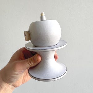 May include: A white ceramic oil lamp with a round body and a cylindrical base. The lamp has a small, round wick and a wooden stopper. The base has a black trim. The lamp is held in a hand.