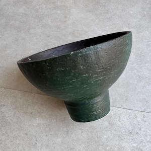 Puede incluir: Un cuenco de cerámica verde oscuro hecho a mano con una forma asimétrica única. El cuenco tiene una superficie texturizada y un interior oscuro. La base es cilíndrica y el cuenco mide unos 20 cm de ancho.