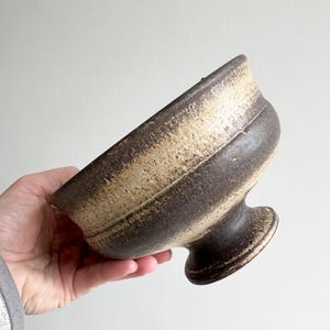 May include: A handcrafted, ceramic bowl with a pedestal base. The bowl features a dark brown exterior with a lighter tan band around the middle. The bowl is held in a hand, showcasing its size and shape. This unique piece is ideal for serving or display.
