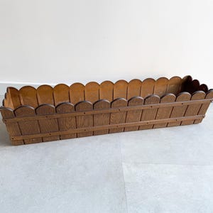 Puede incluir: Una maceta de madera marrón con un borde superior festoneado. La maceta está diseñada para parecerse a una valla de estacas.