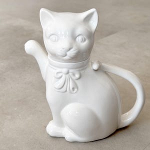 May include: A white ceramic teapot in the shape of a cat. The teapot has a handle, a spout, and a lid. The cat has a bow around its neck and is sitting upright.
