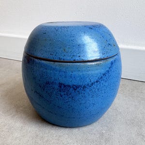 Pode incluir: Um pote de cerâmica azul com tampa. O pote tem formato arredondado e textura salpicada. A tampa se encaixa perfeitamente. O pote é de um tom azul vibrante, com detalhes em azul escuro. O pote está sobre uma superfície cinza claro.