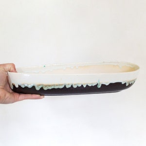 May include: A white oval ceramic planter with a brown and teal glaze. The planter has a drip glaze effect around the rim.