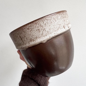 May include: A brown ceramic planter with a textured, off-white rim. The planter has a rounded shape and is held against a white background. The dark brown color contrasts with the lighter rim.