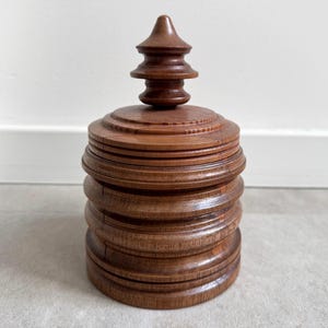 Pu&ograve; includere: Una scatola di legno con un design a strati, a pi&ugrave; livelli e un coperchio appuntito a pi&ugrave; livelli. La scatola &egrave; di colore marrone scuro e ha una finitura liscia e lucida.