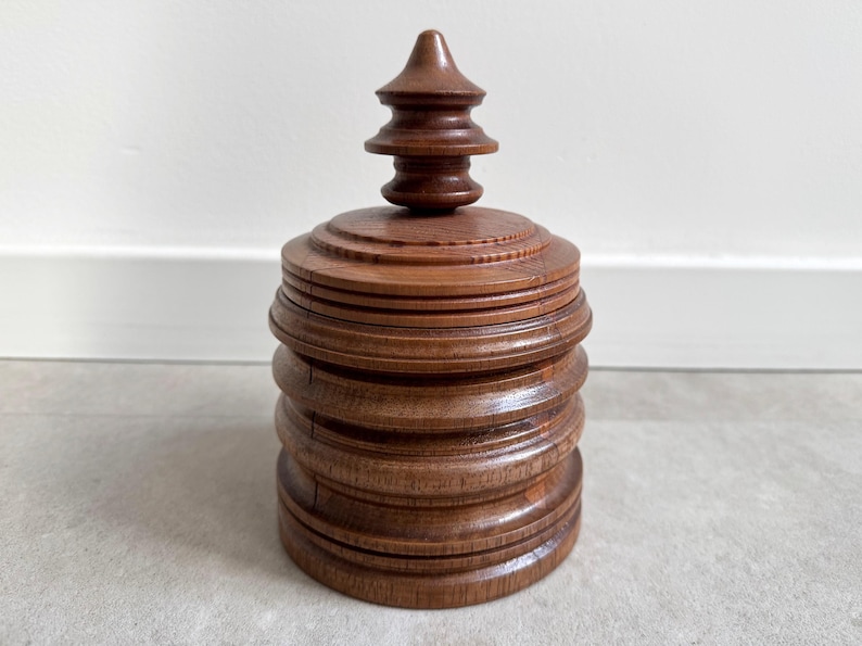 Pu&ograve; includere: Una scatola di legno con un design a strati, a pi&ugrave; livelli e un coperchio appuntito a pi&ugrave; livelli. La scatola &egrave; di colore marrone scuro e ha una finitura liscia e lucida.