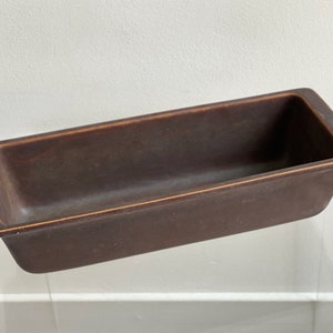 Puede incluir: Un molde para pan de cerámica marrón con un borde ligeramente elevado. El molde tiene un acabado esmaltado y es de forma rectangular.