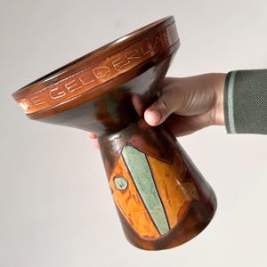 Op de afbeelding: Een bruine keramische vaas met een uitlopende bovenkant en een smallere basis. De vaas heeft een geometrisch ontwerp in oranje en groen. De woorden "DE GELDERLAN" zijn rond de bovenrand gegraveerd. De vaas wordt door een persoon vastgehouden.