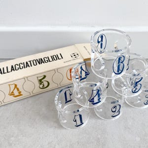 May include: Clear acrylic napkin rings with navy blue numbers 1 through 6 stacked in a pyramid. A box with the text "6 ALLACCIATOVAGLIOLI" is next to the rings. The numbers are in a classic font.