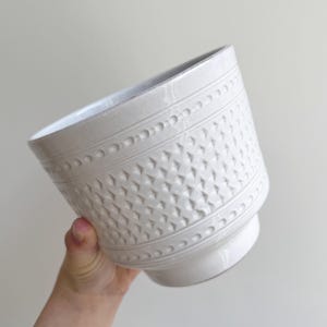 Op de afbeelding: Witte keramische bloempot met een textuur. De bloempot heeft een herhalend geometrisch patroon en een decoratieve band met kleine, verhoogde stippen. De bloempot wordt onder een hoek gehouden, wat het ontwerp en de vorm laat zien.