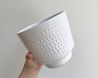 以下が含まれることがあります： テクスチャ加工された表面を持つ白いセラミック植木鉢。植木鉢は、繰り返しの幾何学模様と、小さな隆起した点の装飾的なバンドが特徴です。植木鉢は角度をつけて保持され、そのデザインと形状を示しています。